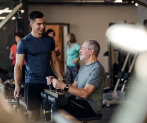 A senior man with a young trainer doing strength workout exercise in gym.